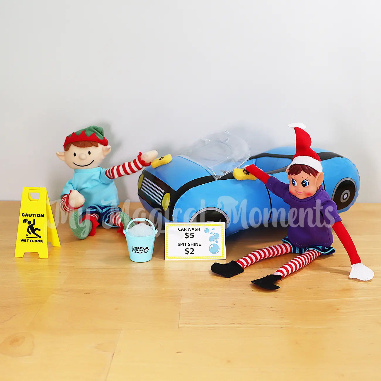 Elves wearing shirts washing an inflatable toy car, a mini wet floor elf sign and bucket with sponges