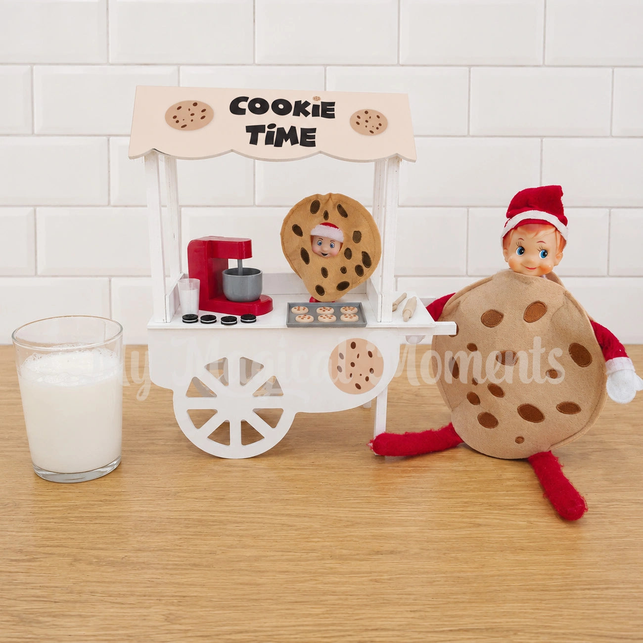 Elf baby dressed in cookie costume with chocolate chips. There is a red baking set, milk with cookies and a tray of baked choc chip cookies Being sold on a cookie stand. There is an elf wearing a chocolate chip cookie costume with a glass of milk