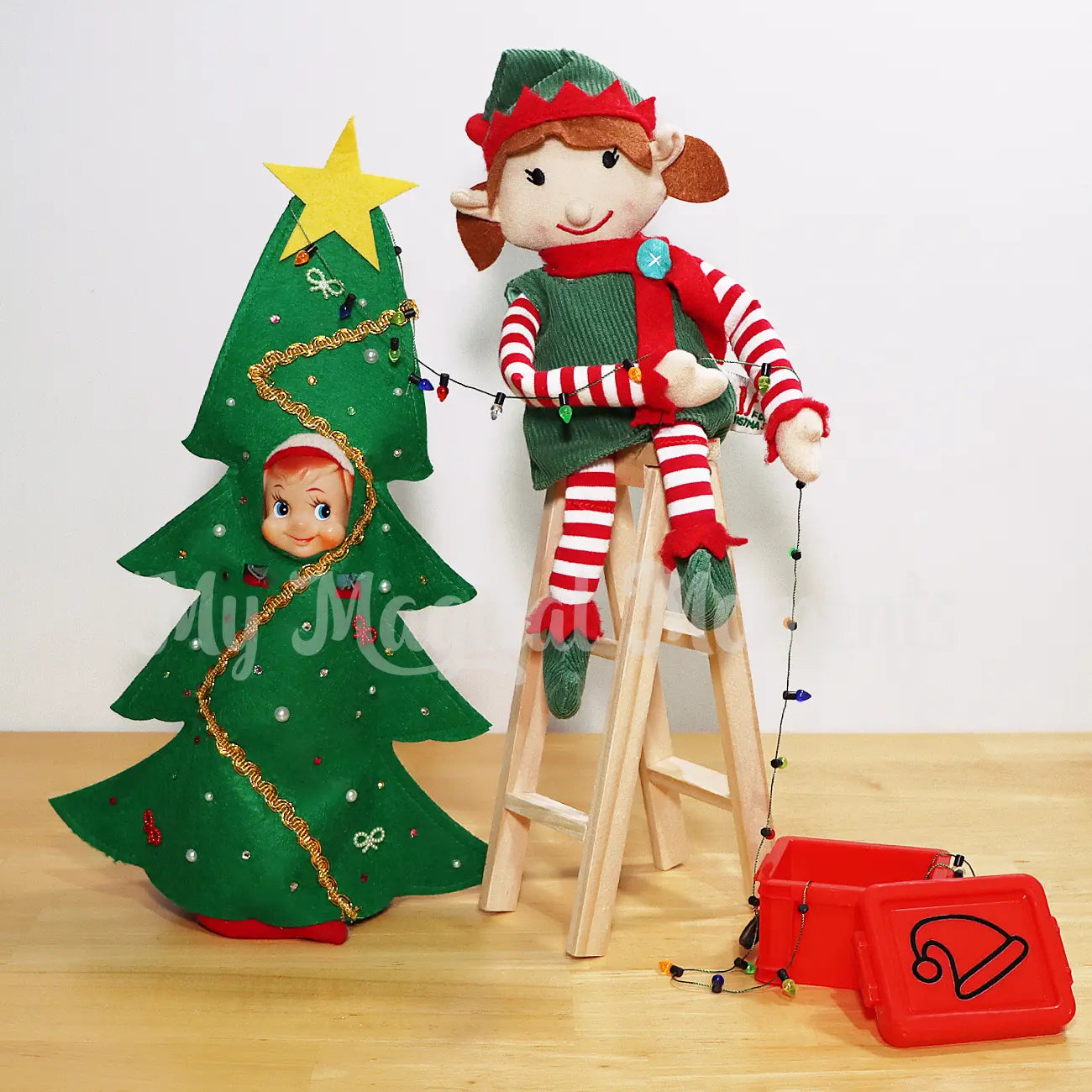 Elf wearing a Christmas tree costume with an elf for Christmas sitting on a ladder holding mini Xmas lights out of a mini container