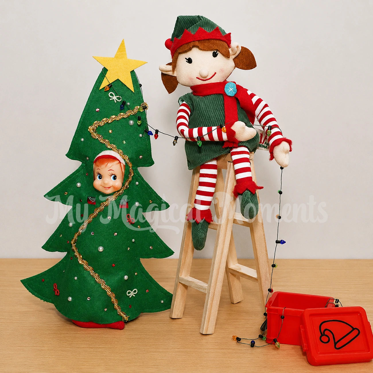 Elf wearing a Christmas tree costume with an elf for Christmas sitting on a ladder holding mini Xmas lights out of a mini container