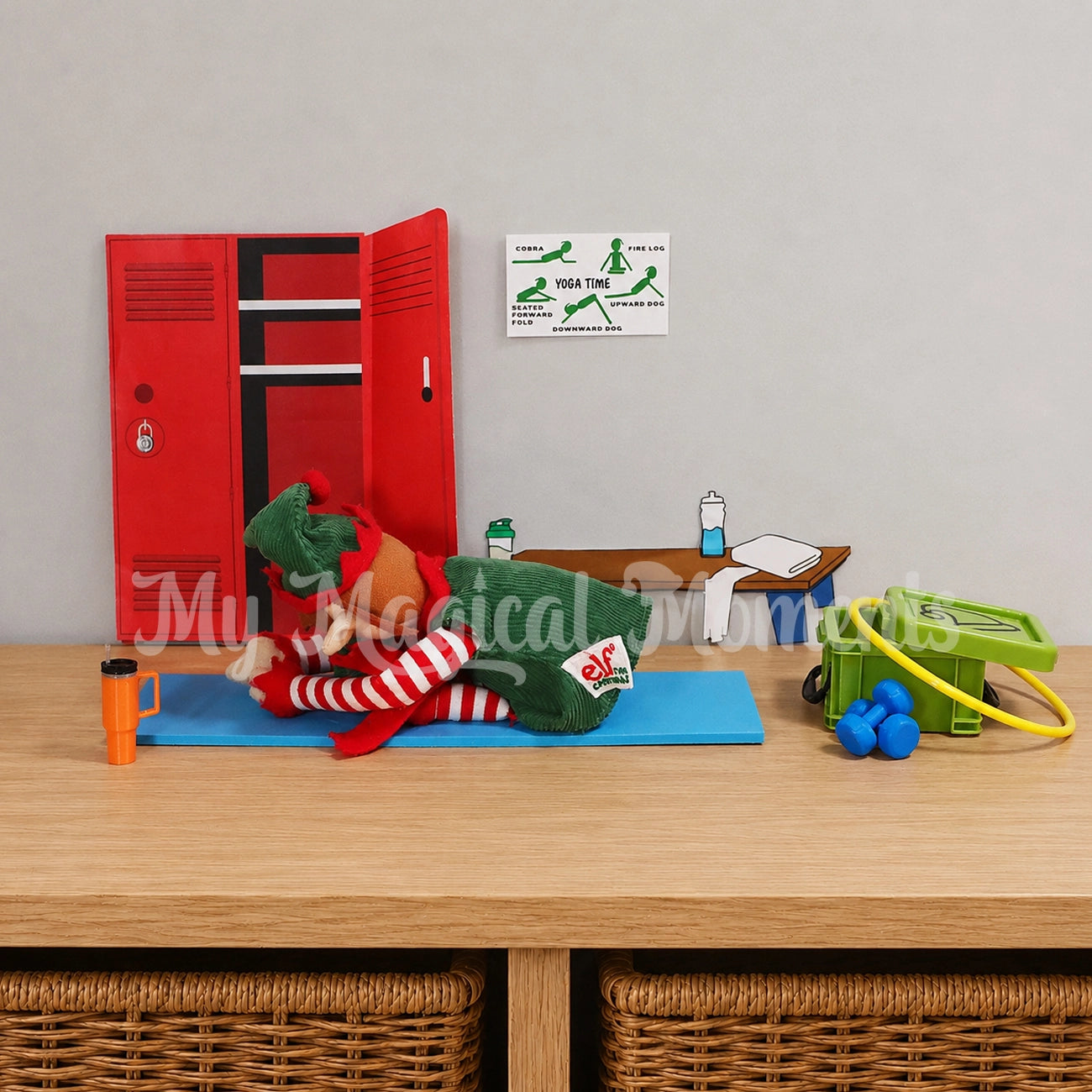 An elf for Christmas doing a yoga pose on a blue yoga mat with a mini elf yoga pose sign. Storage container full of gym equipment, hula hoop and dumbbells. With a stanley cup