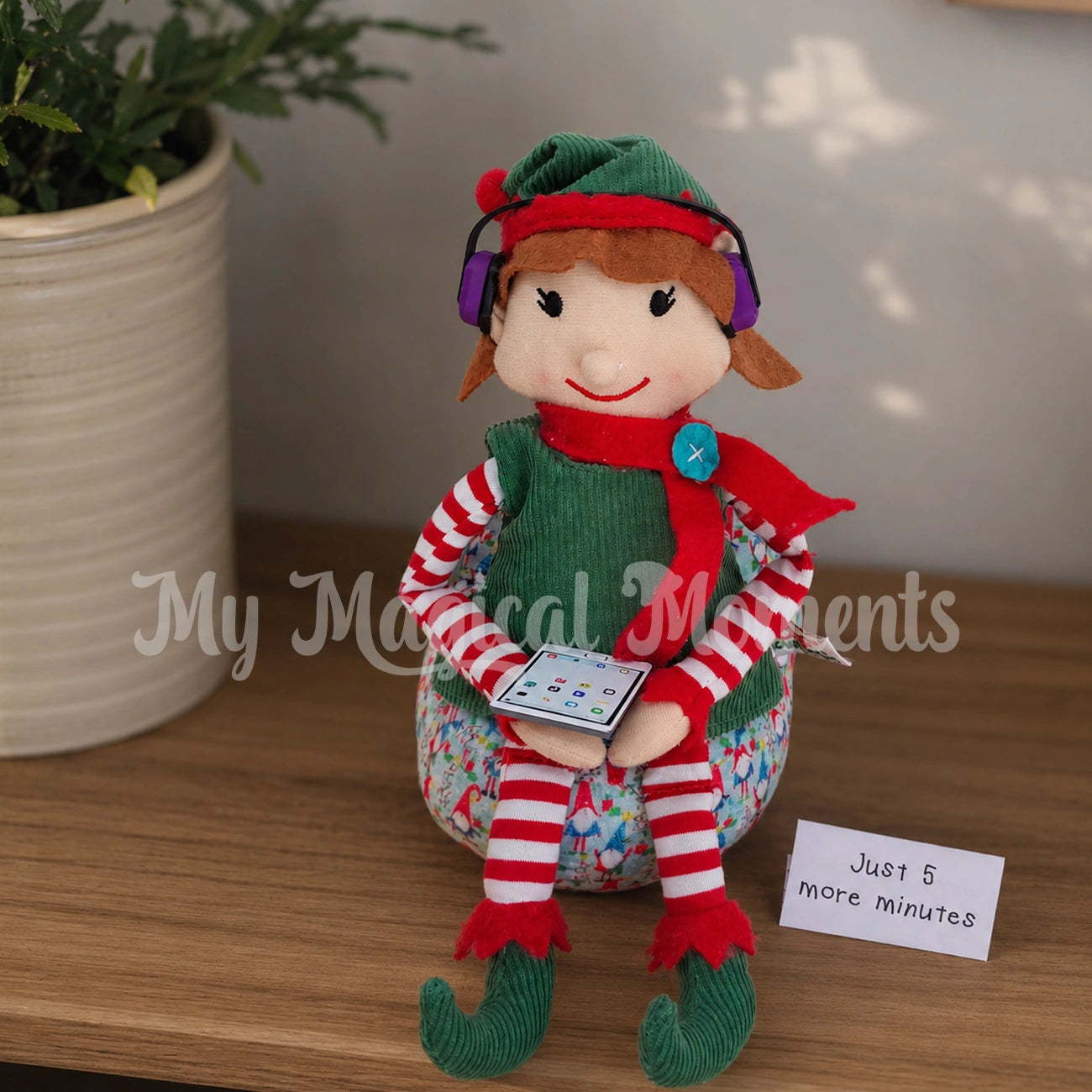 elf playing on a mini ipad tablet sitting on a ban bag with gnomes and wearing purple head phones on a bench. The sign reads just 5 more minutes
