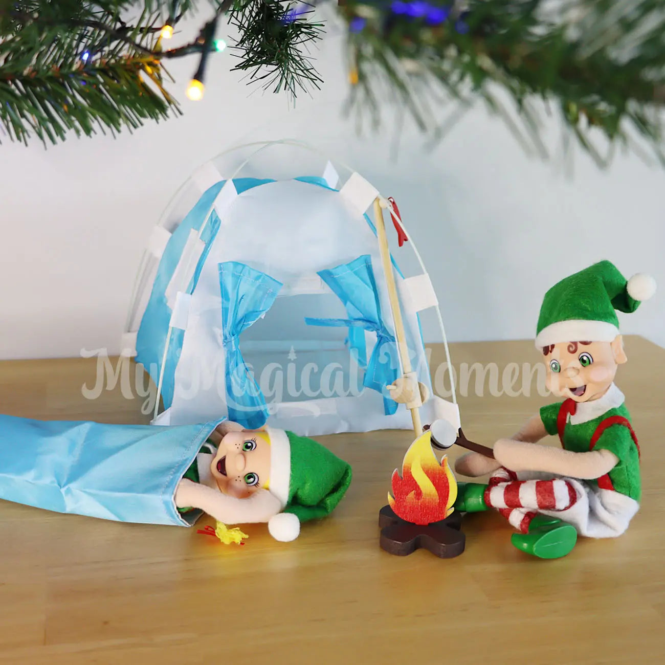 Elves camping under the Christmas tree with mini elf tent, fishing rod and sleeping bag. One elf is toasting marshmallows
