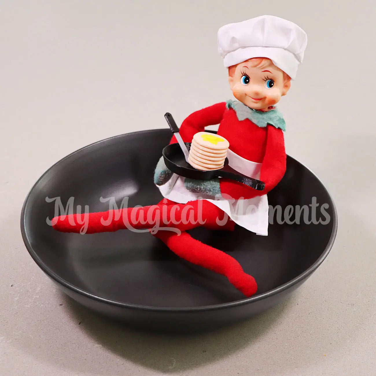 Elf on the Shelf holding a frying pan with pancakes in a black bowl. wearing a chef costume