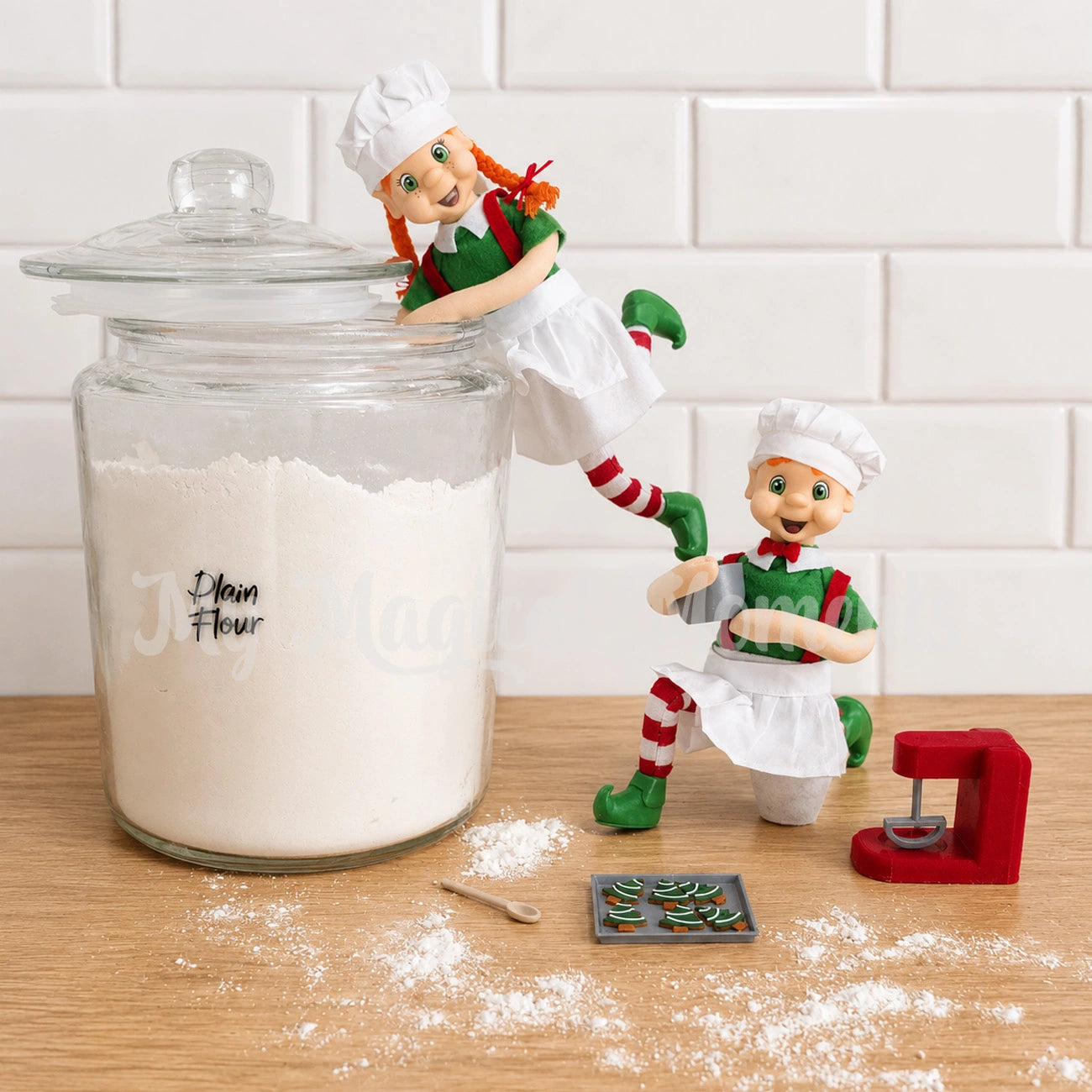 Elf With mini tray of gingerbread Tree cookies and baking set wearing a chef outfit, whilst a second shelf is getting scoops of flour out of a giant glass jar