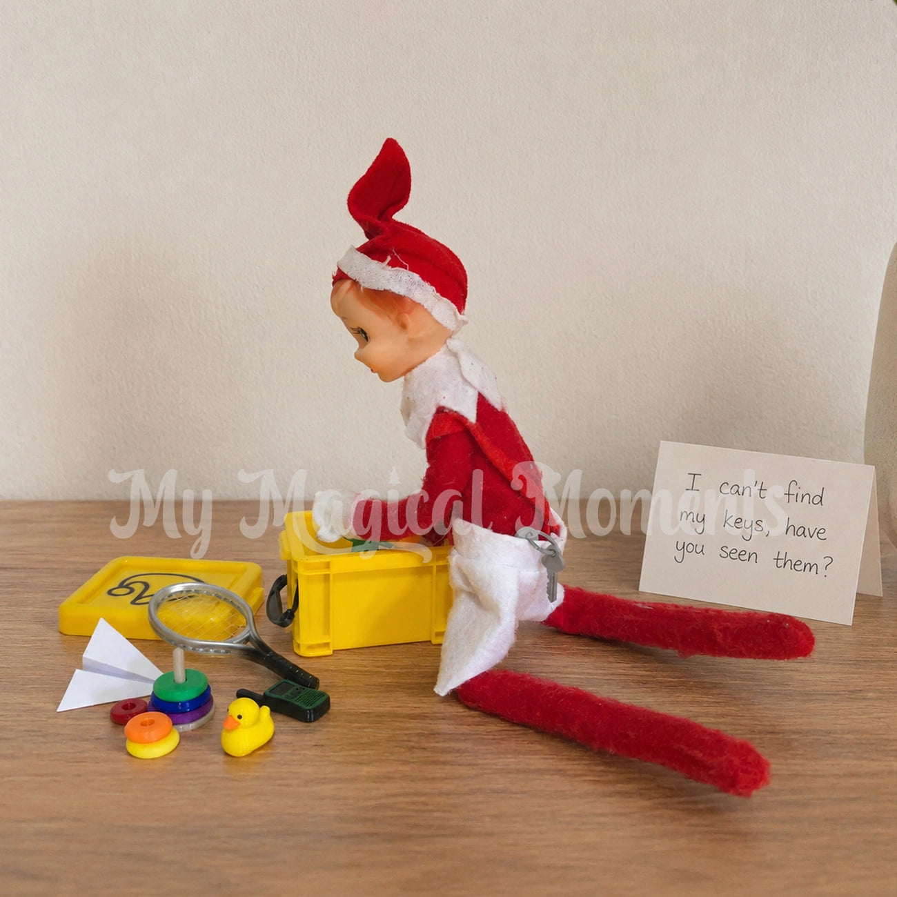 elf looking for their mini house keys in amongst a container of toys, rubber duck, walkie talkie, ring stacker, tennis racquet and paper airplane. There is a note that says I cant find my keys, have you seen them?