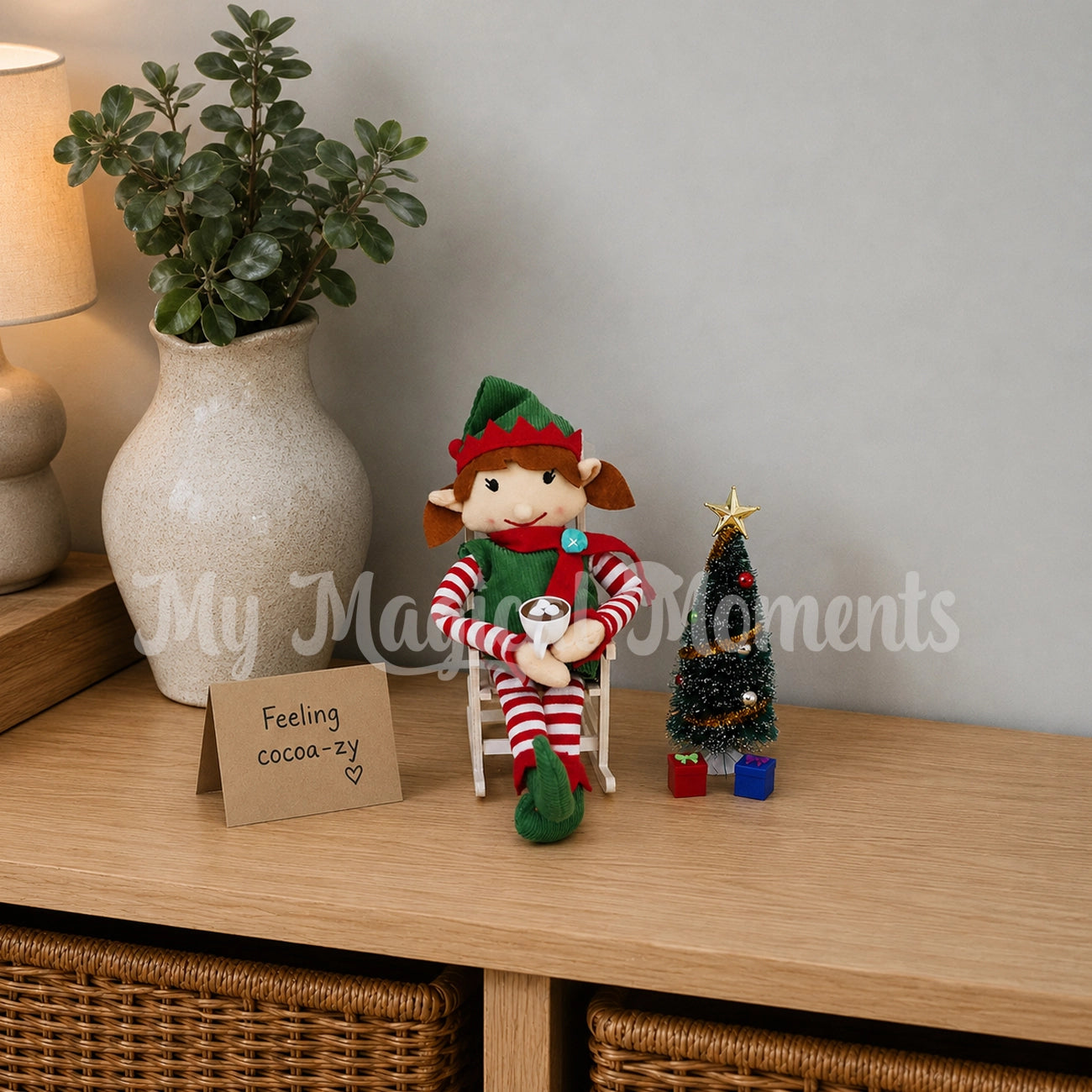 Elf in a wooden rocking chair drinking hot chocolate next to a Christmas tree with mini presents
