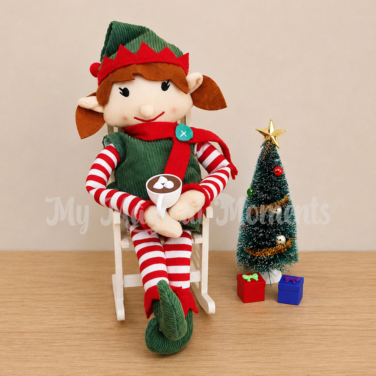 Elf in a wooden rocking chair drinking hot chocolate next to a Christmas tree with mini presents
