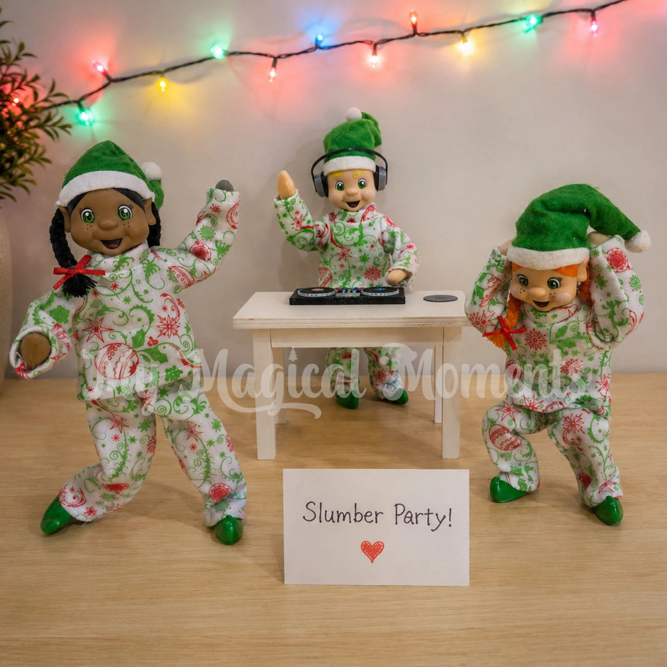 Three bendable standing elf dolls dressed in green and white holiday pajamas pose around a small table With an elf playing a mini DJ Turntable. with a sign reading 'Slumber Party!'. Colorful string lights hang in the background