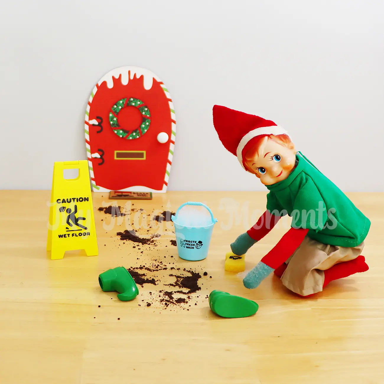 Elf shelf wearing a green shirt cleaning dirty footprints left from shoes, a wet floor sign is out the front of the elf door and door mat. Elf is using a bucket and sponge