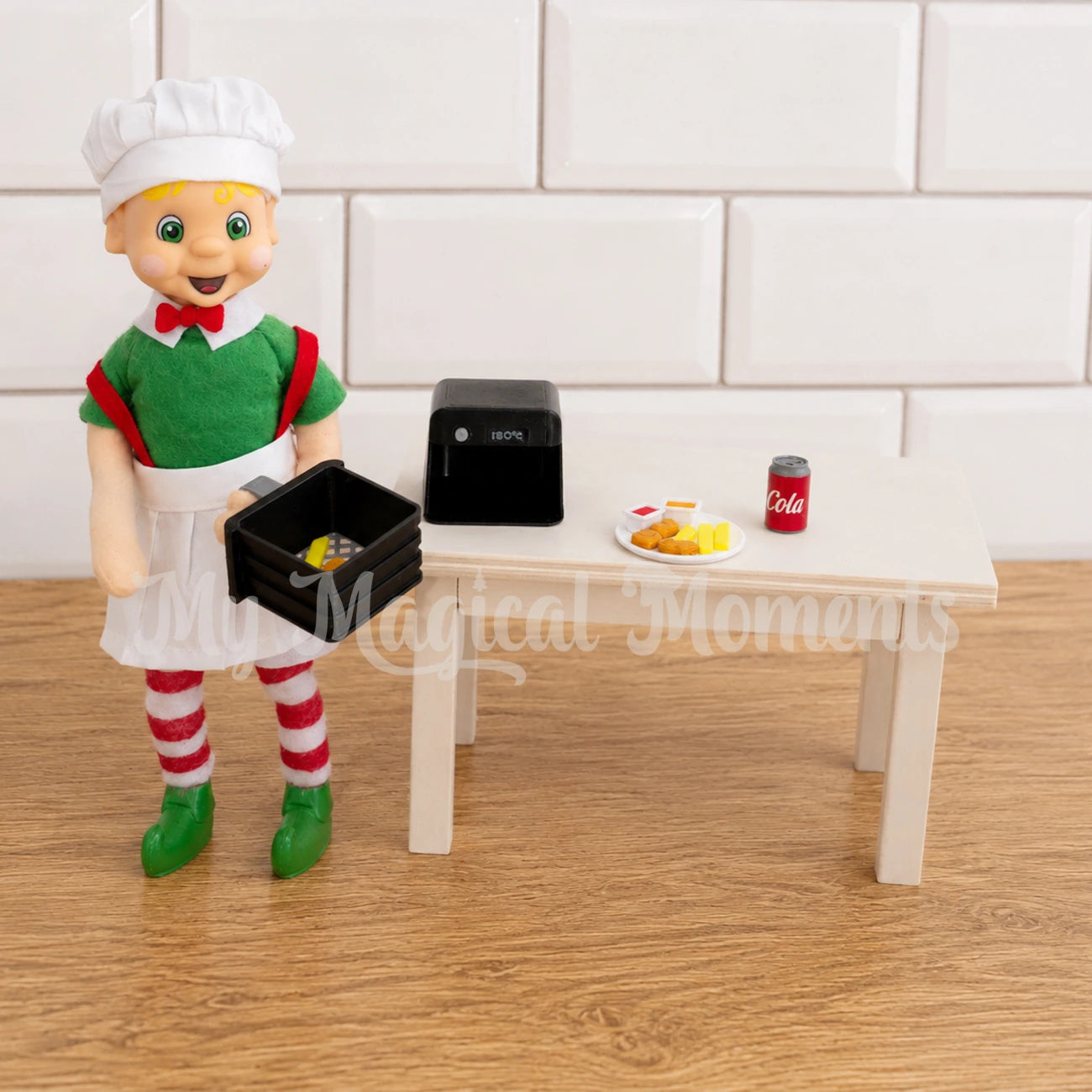 Bendable standing blonde hair elf, wearing a chef costume, holding a mini air fryer tray with chops and nuggets in it, he has a wooden table behind him. On the table there is a plate of mini nuggets, sauce and chips. and a can of cola