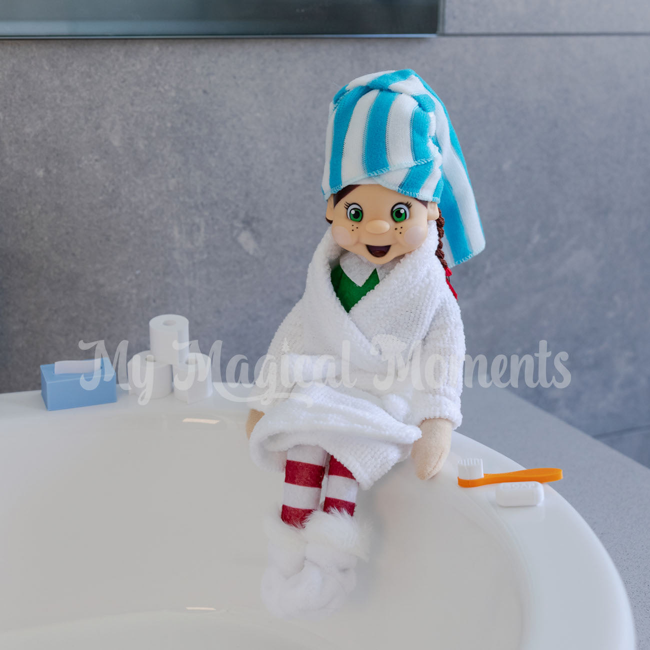 Elf wearing a bathrobe and a mini towel sitting on the sink with mini toilet paper and toothbrush