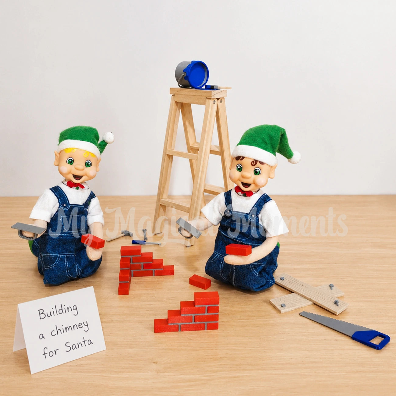 Blonde hair bendable elf wearing overalls building a chimney out of bricks, there is a brown hair boy elf doing the same. There is tools, mini plants of wood, a wooden ladder and a paint tin. There is a folded sign that says Building a chimney for Santa