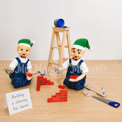 Blonde hair bendable elf wearing overalls building a chimney out of bricks, there is a brown hair boy elf doing the same. There is tools, mini plants of wood, a wooden ladder and a paint tin. There is a folded sign that says Building a chimney for Santa