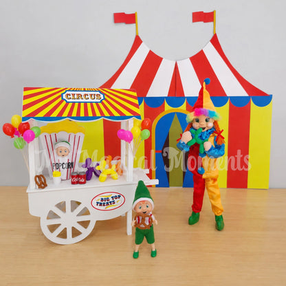 Orange hair elf wearing a clown costume, in front of a printable circus tent, a cart selling mini toy poodle balloons, miniature cola cans, fries and pretzels. There is a baby elf dressed as a popcorn and balloons. The toddler elf is eating a pretzel