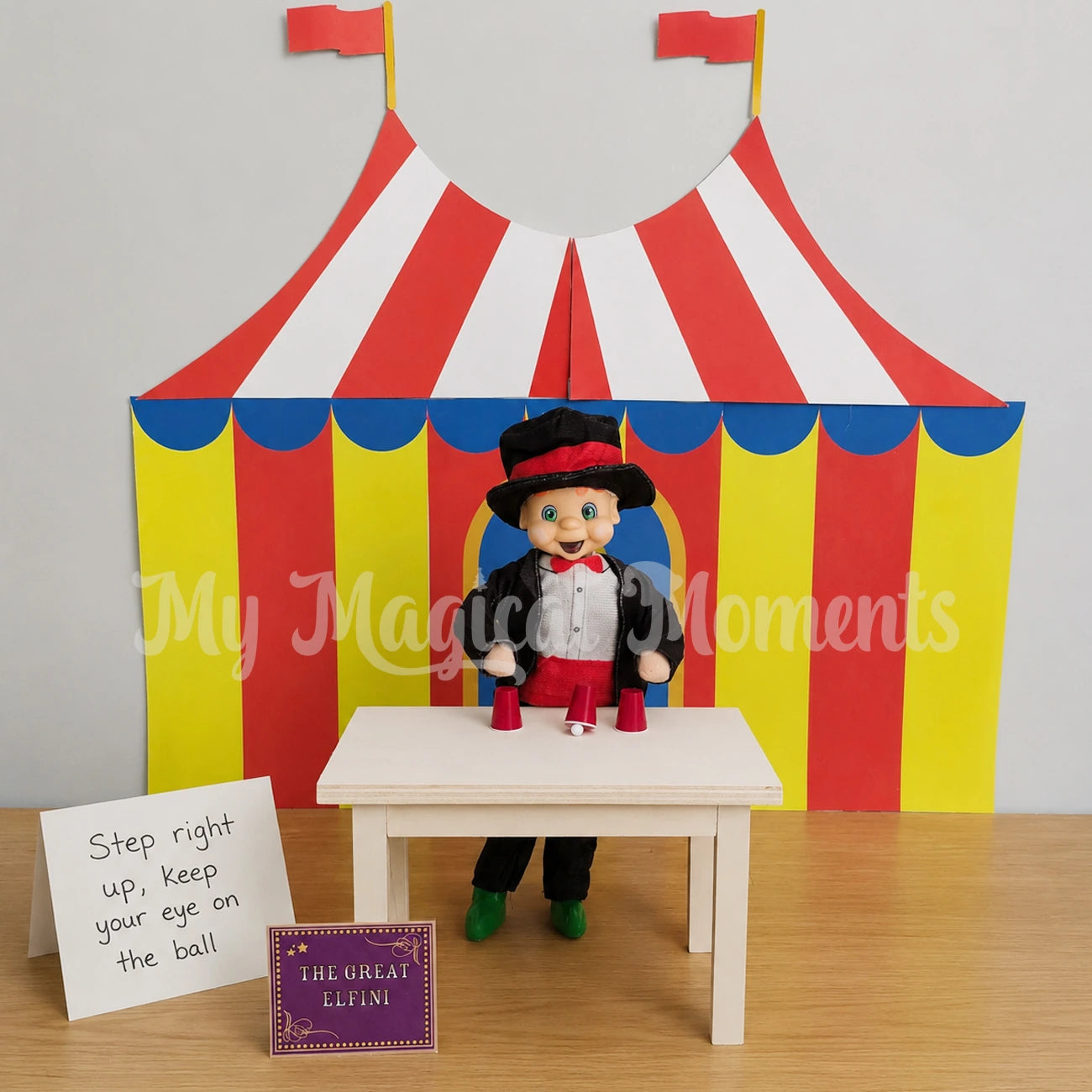 Elf dressed as a magician doing a magic cup trick in front a printable circus tent. Using a wooden mini table with a sign that says step right up, keep your eye on the ball as the great elfini