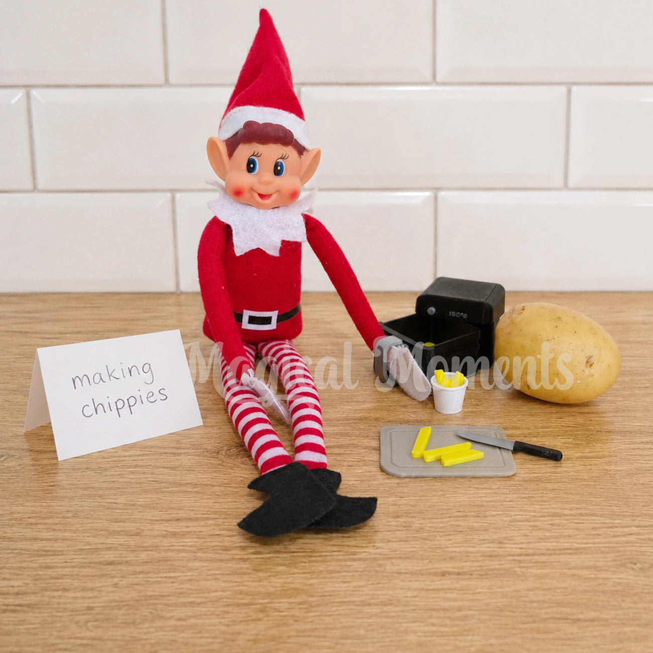 Elf holding an air fryer chopping a potato to make chips with a mini chopping board and knife set. With a sign saying making chippies