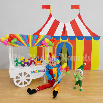 Elf wearing a clown costume, holding balloons selling mini balloon animals at a Balloon stand in front of a circus tent. There is a toddler elf holding a balloon animal