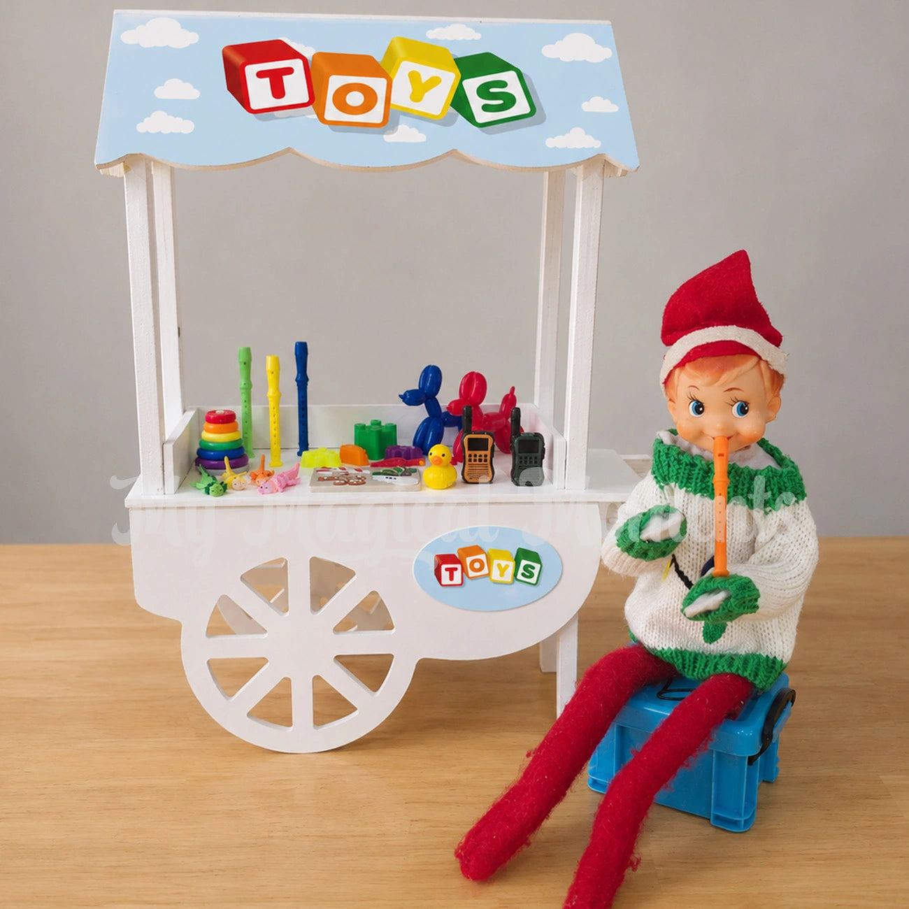 Elf wearing an ugly Christmas sweater playing an orange recorder, sitting on a elf container. Selling toys on  a stand, their is a little ring stacker, mini fidget toys, rubber ducky, mini beach toys, miniature walkie talkies and balloon animals