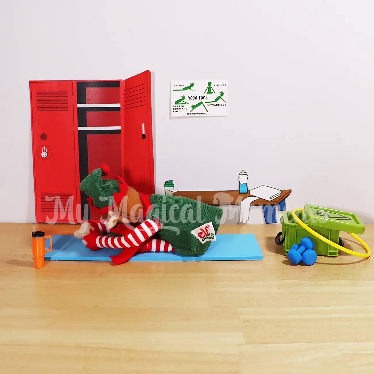 An elf for Christmas doing a yoga pose on a blue yoga mat with a mini elf yoga pose sign. Storage container full of gym equipment, hula hoop and dumbbells. With a stanley cup