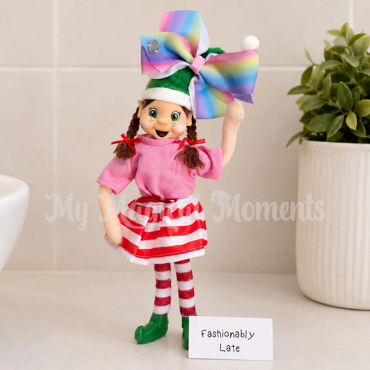 Elf wearing a red & white skirt, with a pink shirt on and a big bow in her hair. the sign reads fashionably late