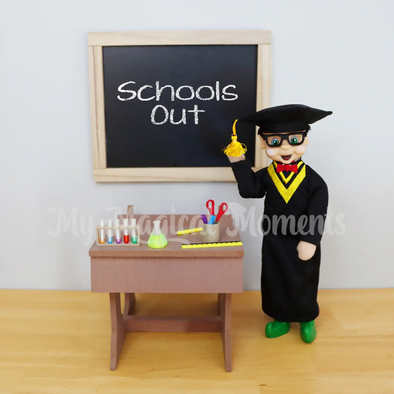 Elf wearing a graduation outfit in front of school desk and mini school supplies