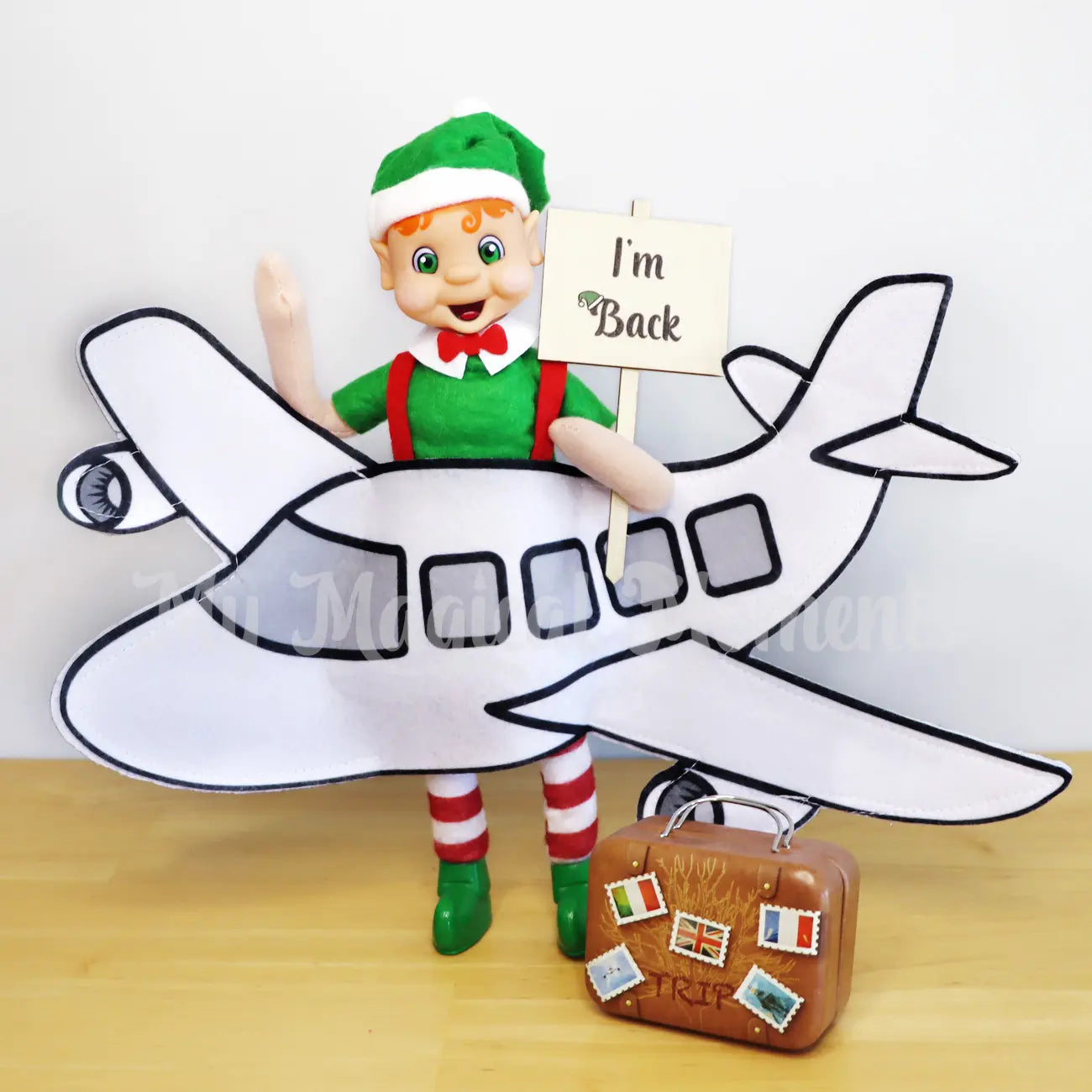 Elf wearing an airplane costume with an I'm back sign and suitcase