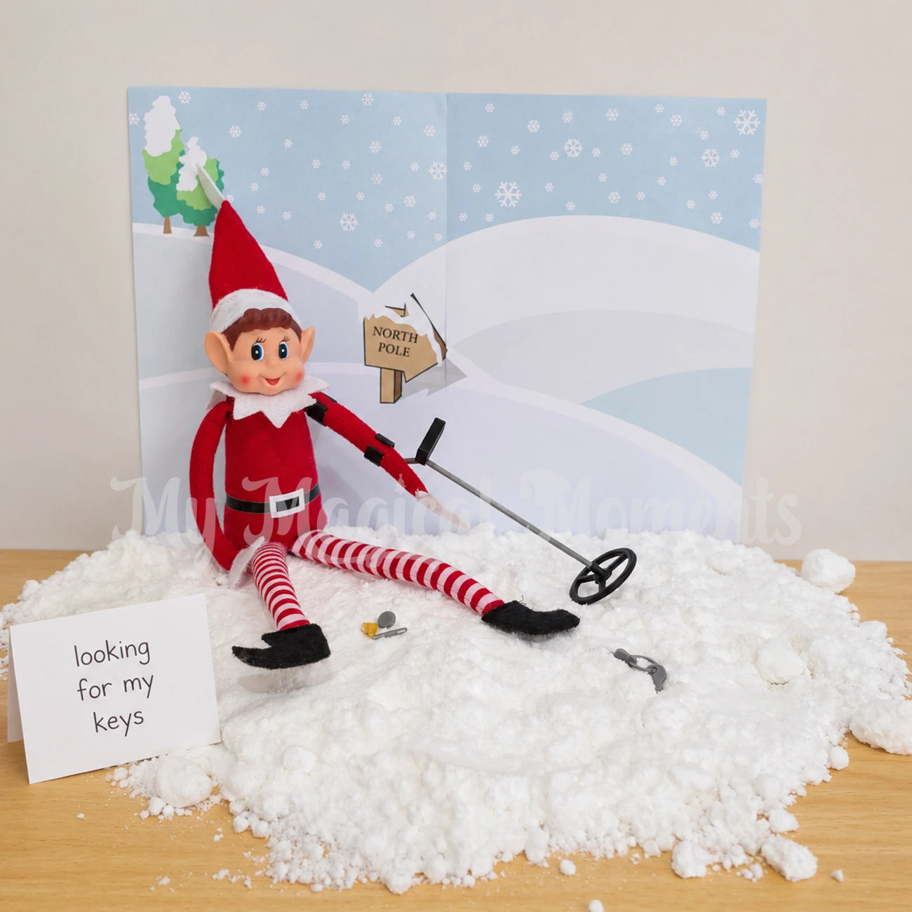 Elf using a mini metal detector to look for his missing keys in the snow, with a mini nugget, paper clip and bottle cap found. Theres a sign saying looking for my keys and a backdrop of a snow scene