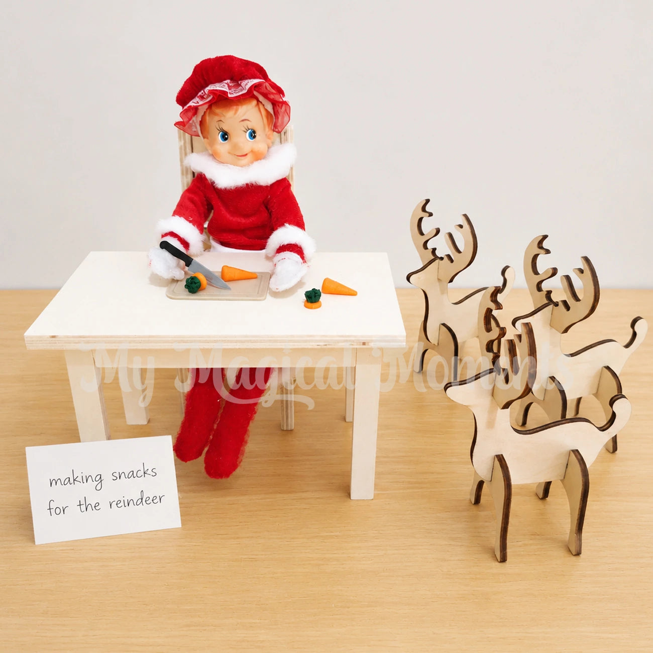 Elf wearing a Mrs Claus costume, cutting up carrots with a mini knife and chopping board set. there are 3 wooden reindeers waiting and a sign leaned against a wooden table that says, making snacks for the reindeer