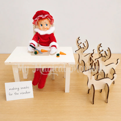 Elf wearing a Mrs Claus costume, cutting up carrots with a mini knife and chopping board set. there are 3 wooden reindeers waiting and a sign leaned against a wooden table that says, making snacks for the reindeer