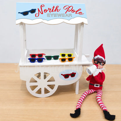 elf wearing glasses, holding a megaphone selling sunglasses from a little shop front