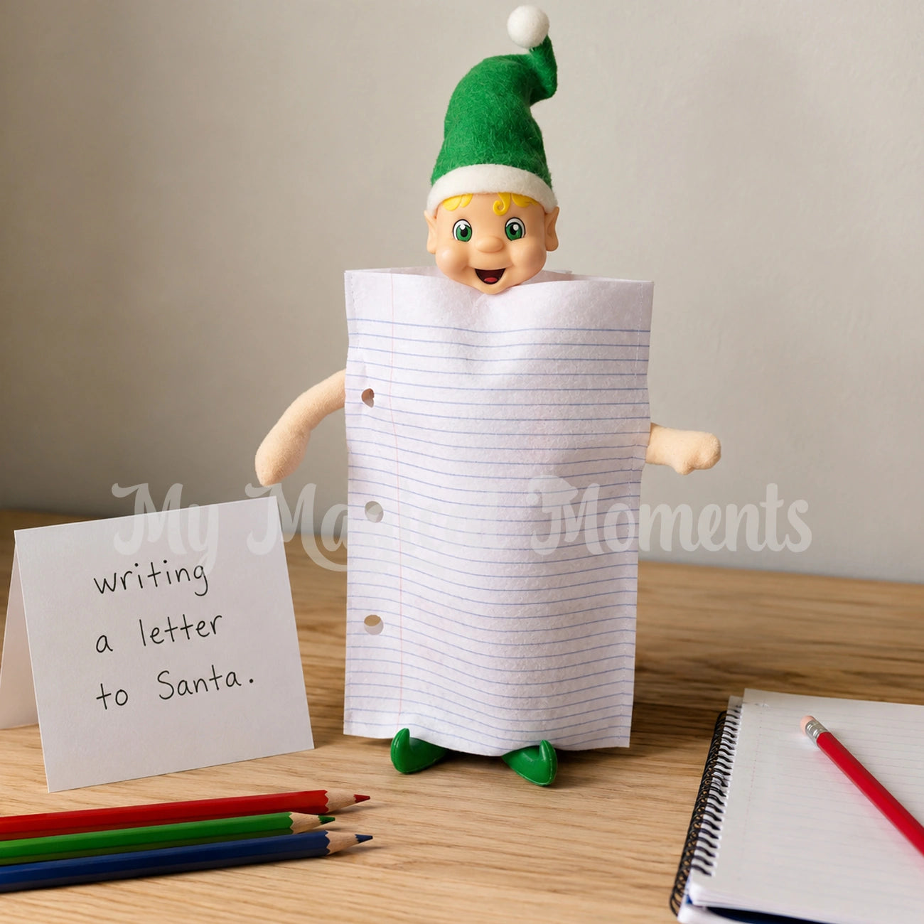 Blonde hair elf dressed in a paper costume, on a table with papers and pencils. There is a sign that says writing a letter to Santa