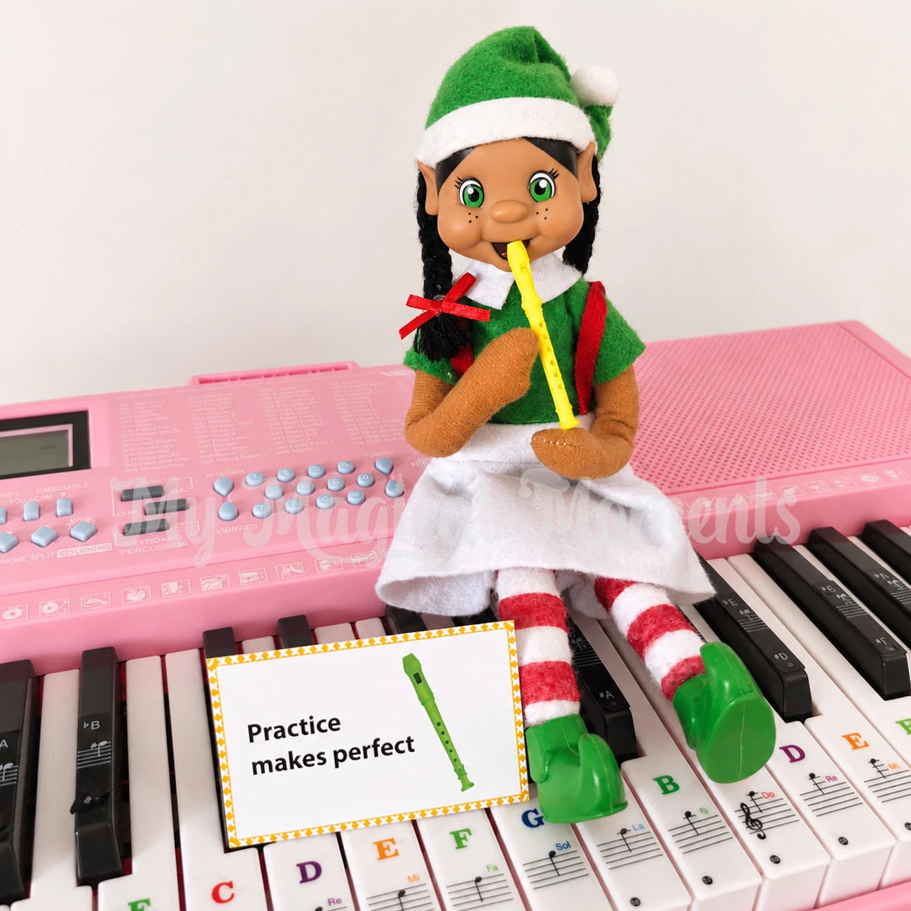 A My elf friends with braided black hair, wearing a green Santa hat, green top, white skirt, and red-and-white striped tights, sitting on a pink toy keyboard and holding a yellow recorder prop. A small sign reads 'Practice makes perfect.