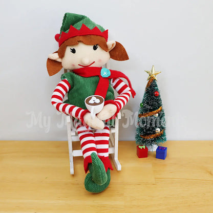 Elf in a wooden rocking chair drinking hot chocolate next to a Christmas tree with mini presents