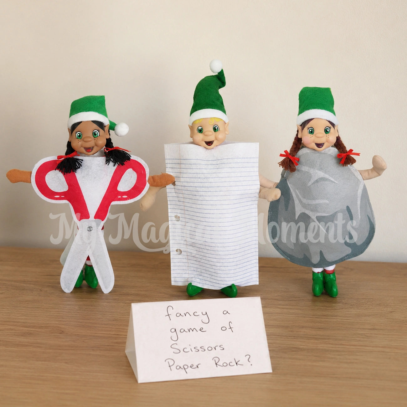 Elves wearing Scissors costume, Paper costume and a rock costume. There is a handwritten sign that says fancy a game of scissors paper rock