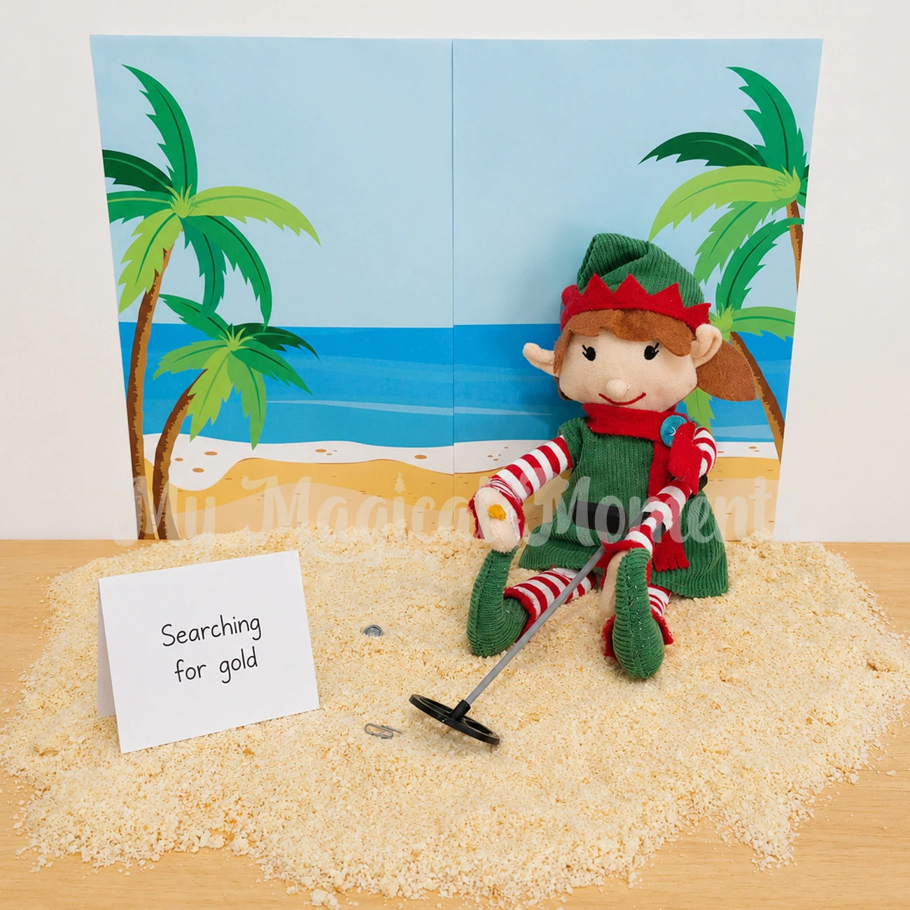 Elf sitting on a pile of bread crumb sand, with a beach background printable. Holding a metal detector looking for gold. The sign says searching for gold