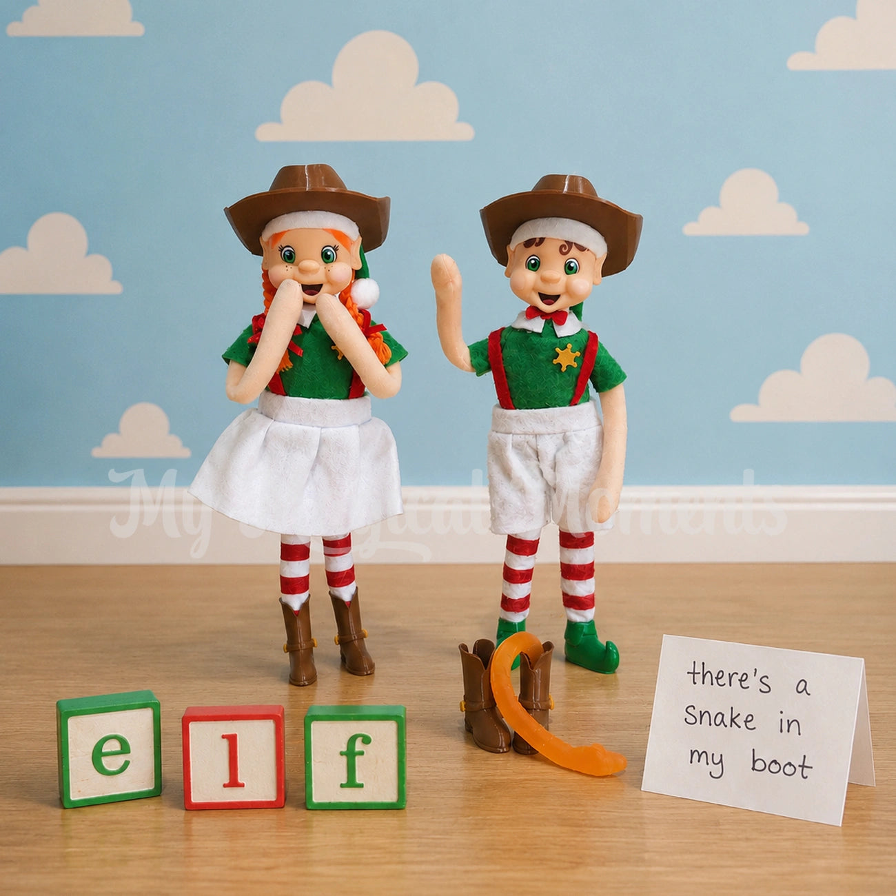 a orange hair bendable elf standing wearing a cowboy hat and boots, a brown hair boy elf standing waving wearing a cowboy hat and sheriff badge, there is two cowboy boots on the ground and a snake coming out of it . the sign says there's a snake in my boot