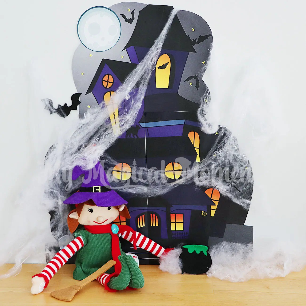 Elf wearing a witch hat, with cauldron, bat and broom stick, in front of a spooky haunted mansion