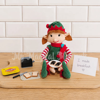 an elf sitting on a  chopping board, with mini toast, vegemite  and a plate full of mite on toast. there is a mini toaster and a note that says I made breakfast