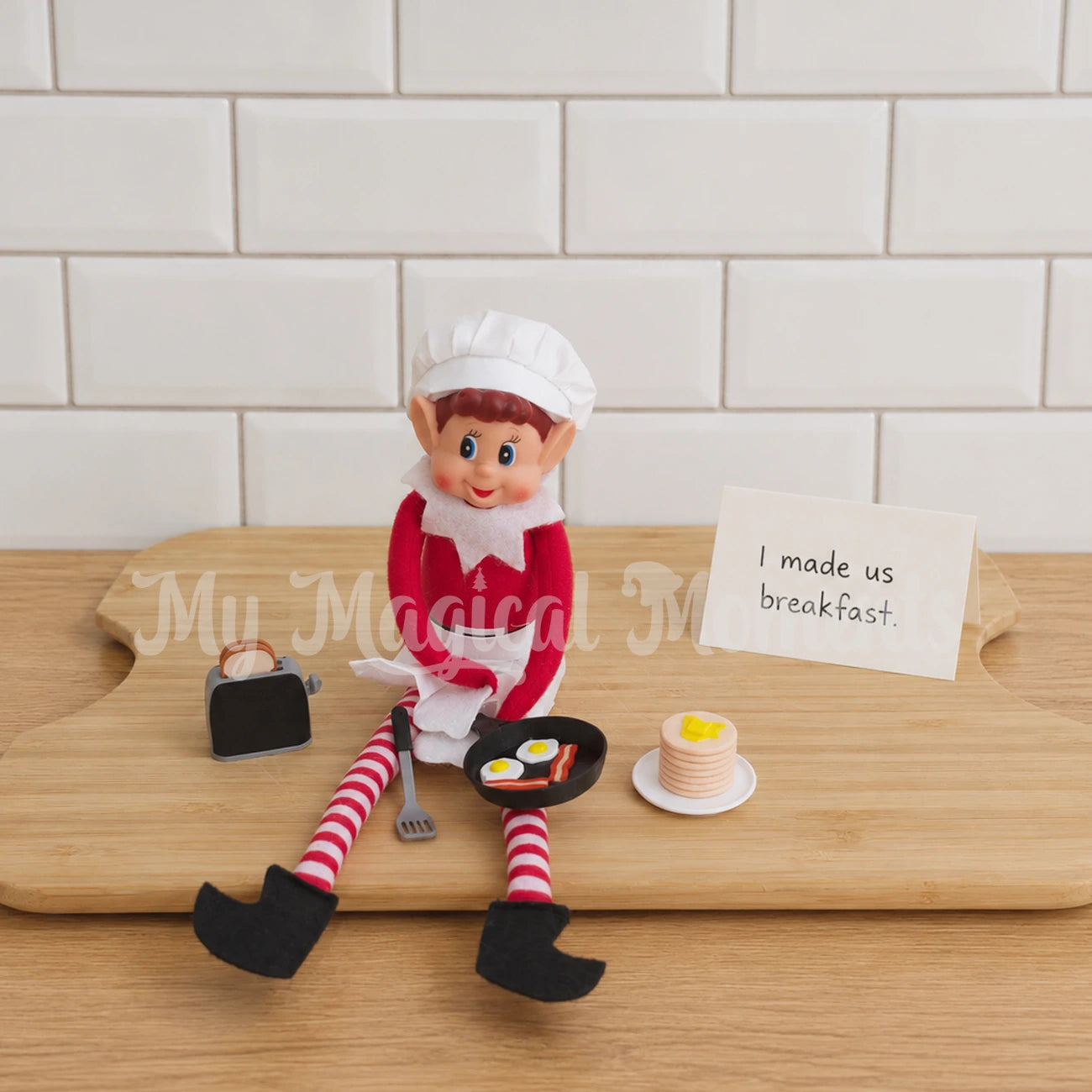 Elf wearing a chef outfit, making breakfast with a mini elf sized toaster, toast, a spatula, mini frying pan and little eggs & bacon in the shape of a smiley face. There is a stack of mini pancakes on a plate with a note that says I made us breakfast