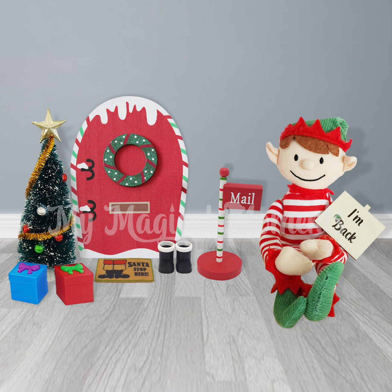 Elf holding an I'm back sign next to elf door and miniature Christmas tree