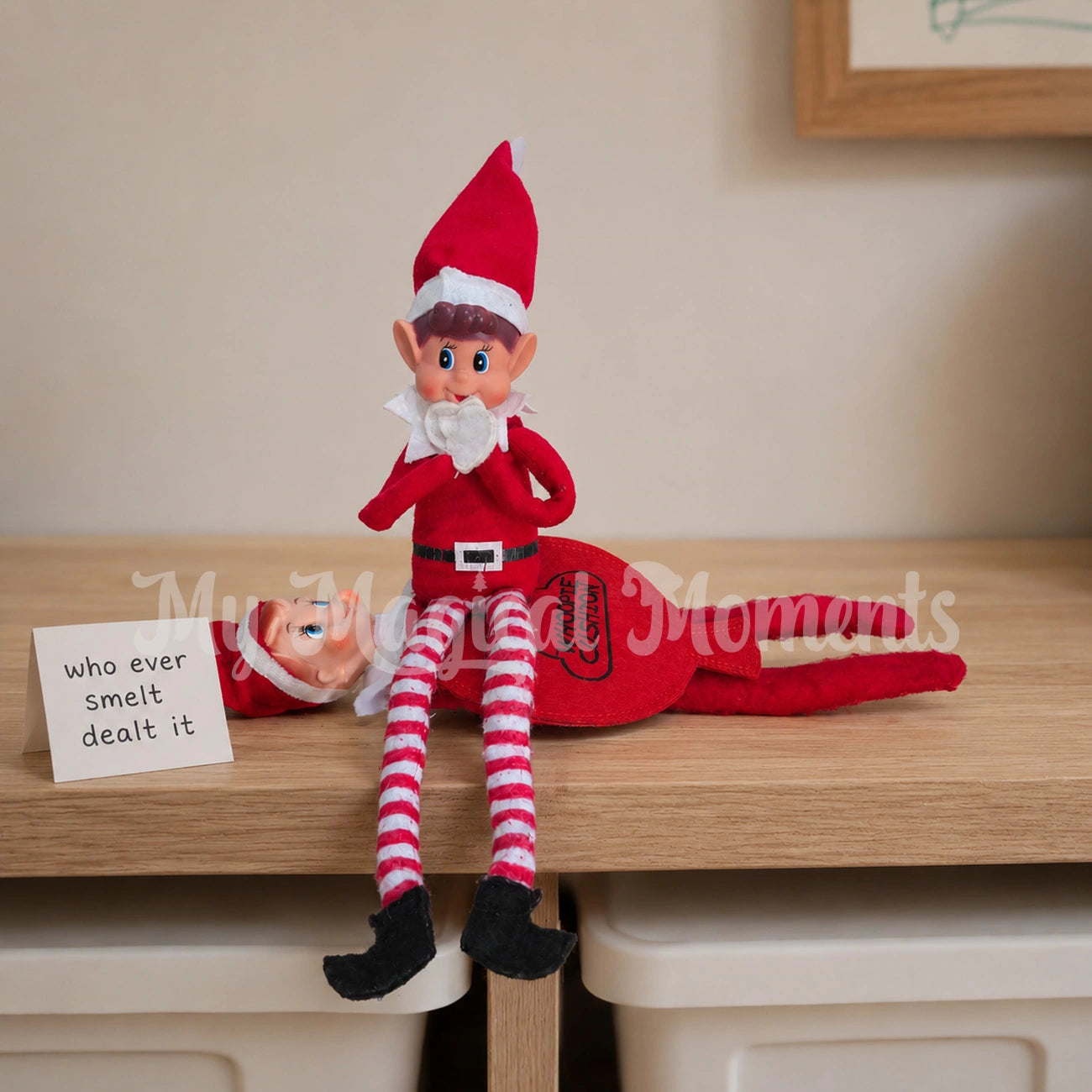 Elf sitting on another elf wearing a whoopie cushion with a sign saying who ever smelt it dealt it