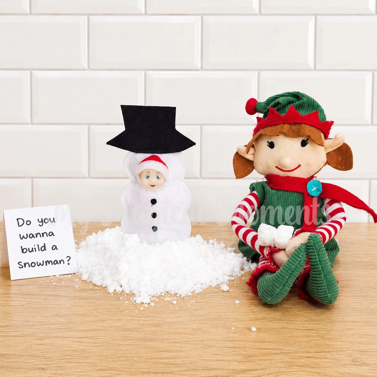 baby elf wearing a snowman costume, surrounded by fake snow and an elf for Christmas in a kitchen with a sign saying do you wanna build a snowman?