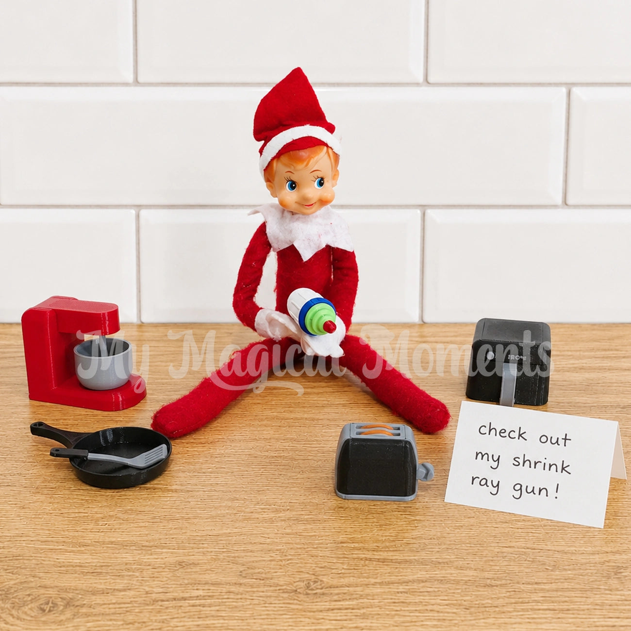 elf in the kitchen using a mini shrink ray to shrink down a bunch of little elf props, a baking set, fry pan and spatula, a toaster and an air fryer. there is a sign that says check out my shrink ray gun