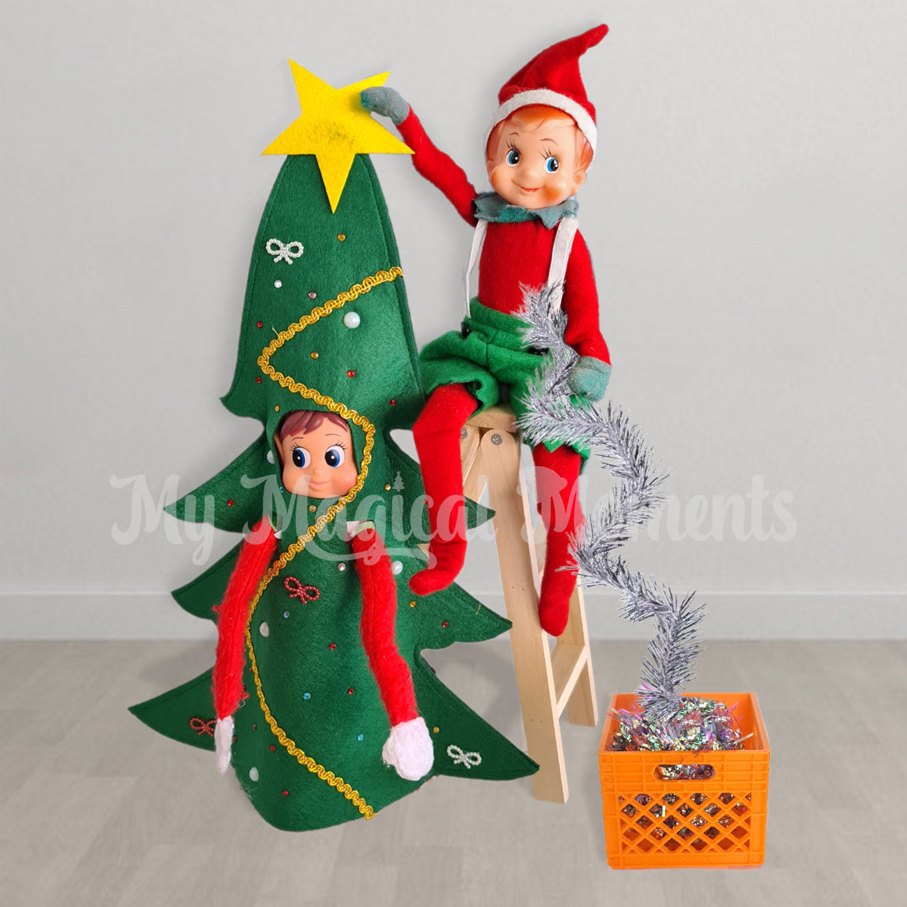 An elf sitting on a ladder decorating another elf dressed as a Christmas tree with a miniature crate full of decorations