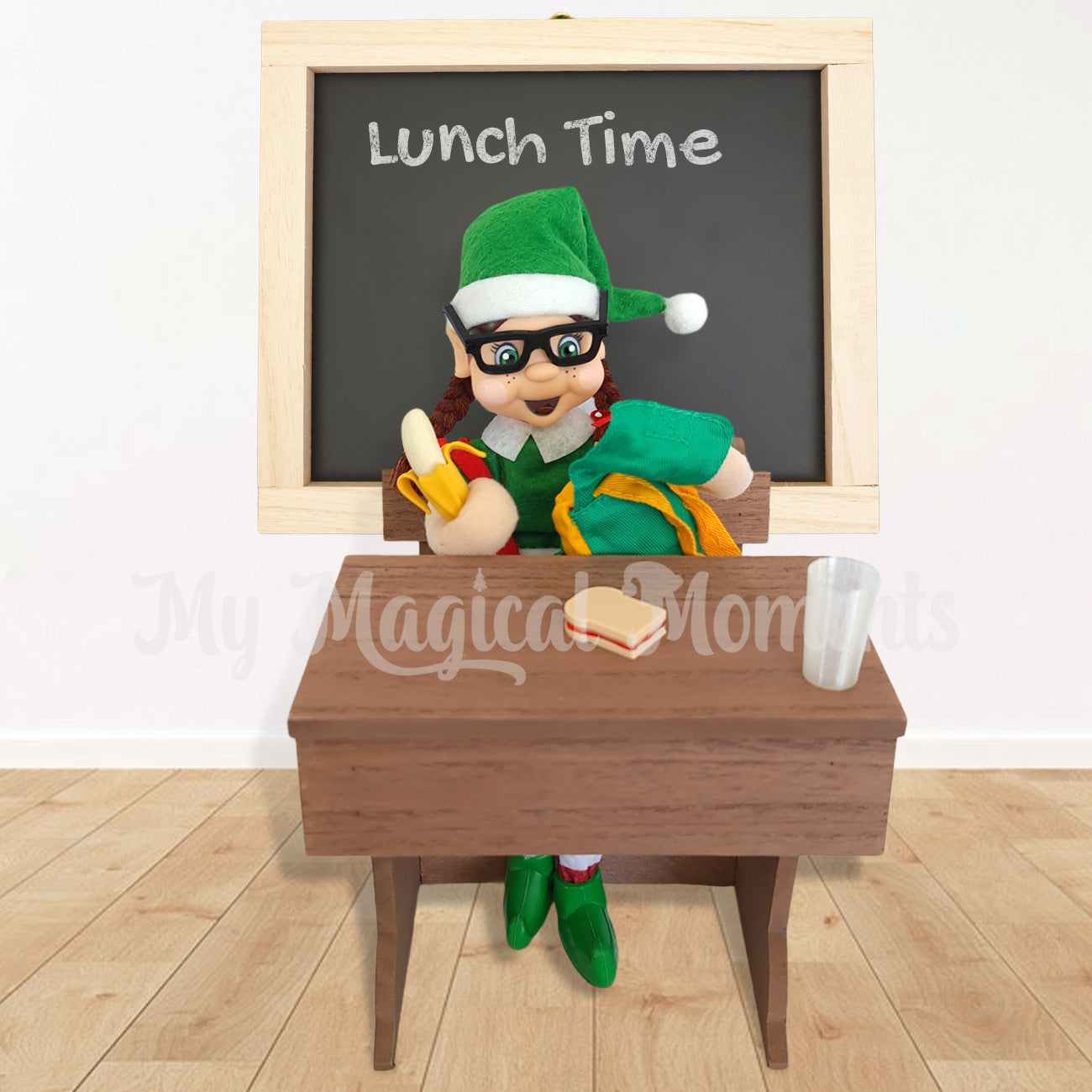 Brown hair elf sitting at a school desk with her miniature backpack and elf props. A chalkboard is behind with the words Lunch time